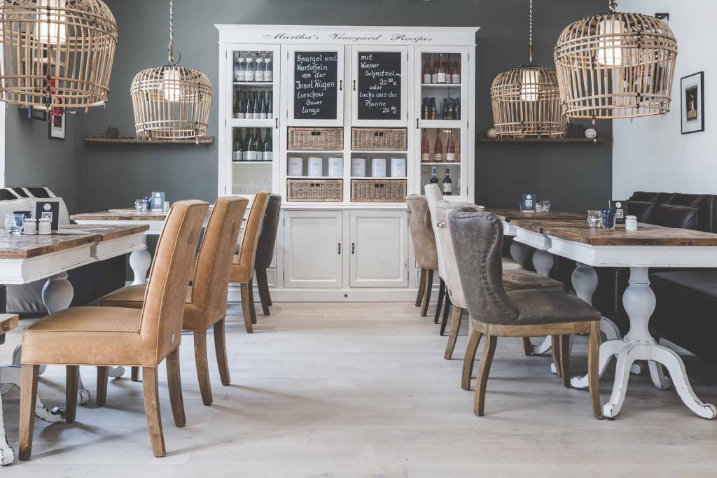 Gemütliches, stilvolles Restaurantinterieur mit Holztischen, hellbraunen und grauen Polsterstühlen, Rattanleuchten und einem weißen Schrank mit Weinflaschen - eines der einladenden Restaurants in Binz mit Speisekarten auf Kreidetafeln an der Rückwand.